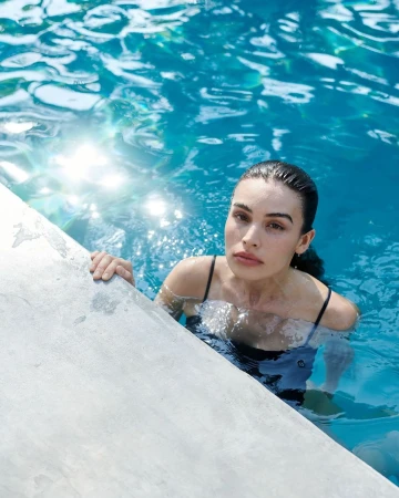 Woman with dark hair and fair skin, partially submerged in a turquoise swimming pool, holds onto the pool's concrete edge, looking directly at the camera.