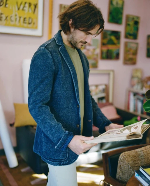Man in a denim blazer examines a book in a room with framed art, a dark chair, and natural light from a window casting soft shadows.