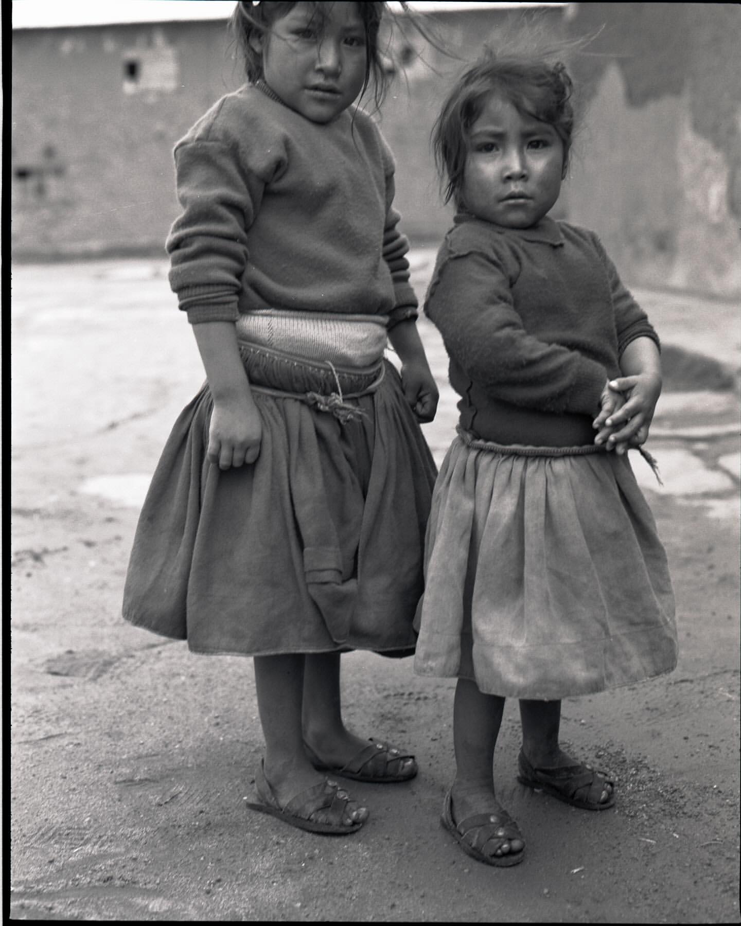 Two young girls stand outdoors in matching outfits, featuring sweaters, skirts, and sandals. The vintage photograph is black and white.