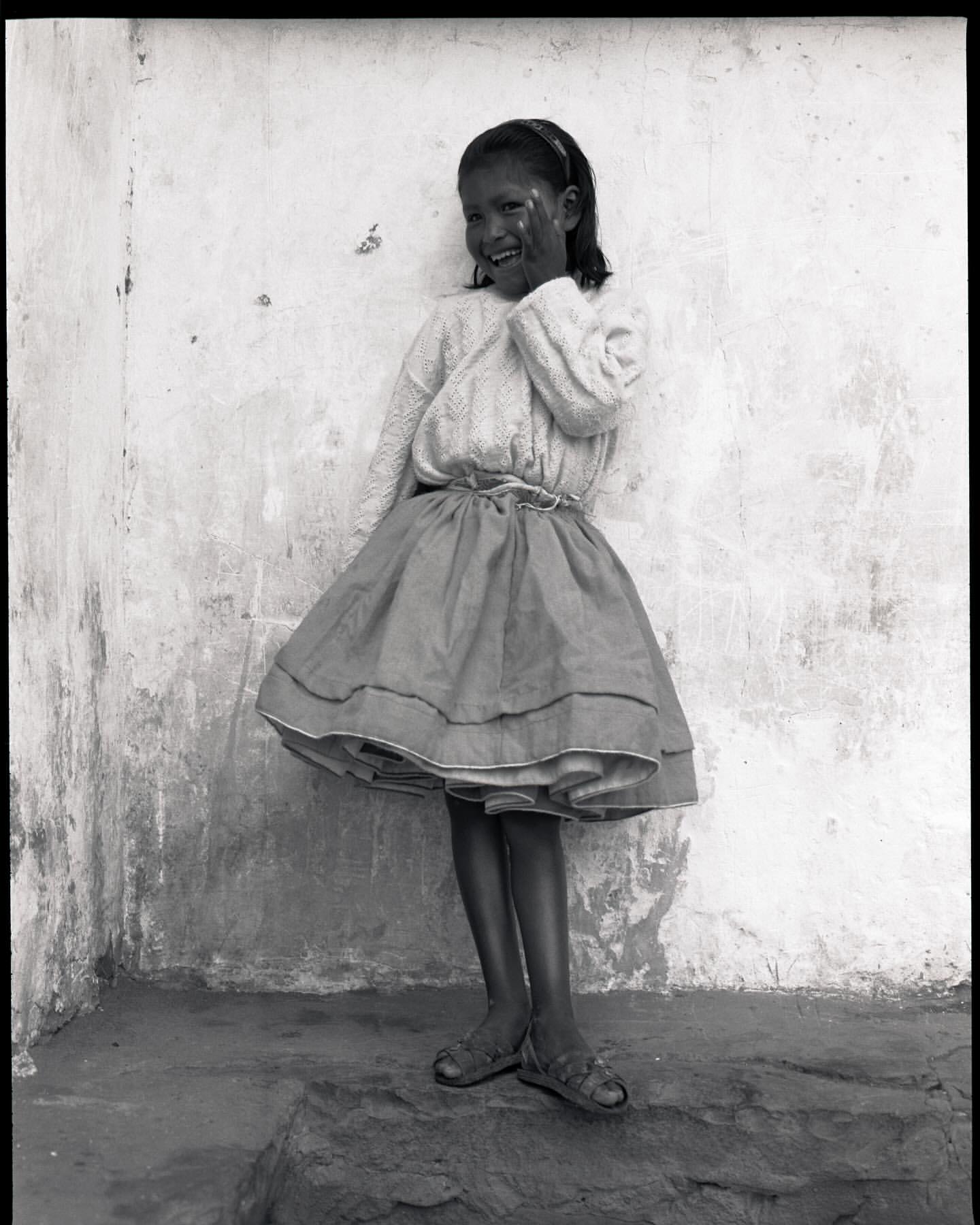 Young girl in dress smiles against weathered wall, captured in black and white medium shot with subtle textures and light.