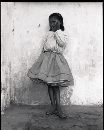 Young girl in dress smiles against weathered wall, captured in black and white medium shot with subtle textures and light.