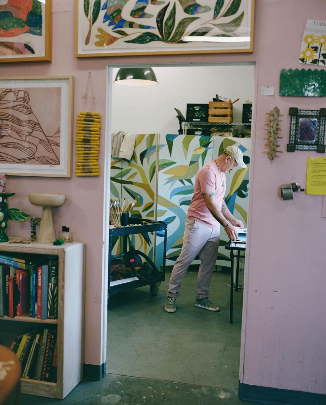 Adult male stands in art studio, pink polo shirt, painting or art creation in progress, framed artworks on wall and equipment visible.