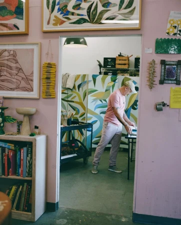 Adult male stands in art studio, pink polo shirt, painting or art creation in progress, framed artworks on wall and equipment visible.