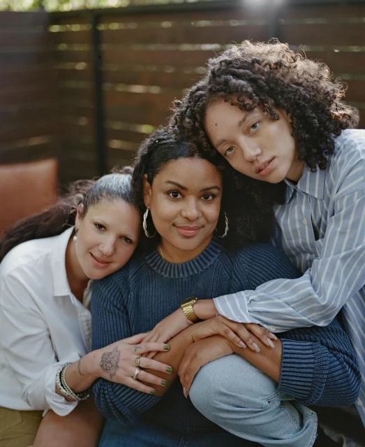Three young women with curly hair and casual outfits are hugging and smiling together in an outdoor setting, portrait shot.