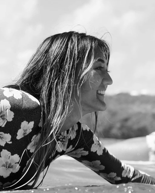 Monochrome portrait of a woman in a floral rash guard, with wet hair, smiling outdoors with a beach and sky backdrop.