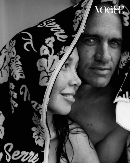 Black and white studio shot of a smiling adult couple embracing under a floral patterned towel, facing forward with visible 'Vogue Living' logo.