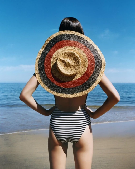 Back view of a woman in a striped swimsuit and a straw hat at the beach, with the blue ocean and sky visible in the background.