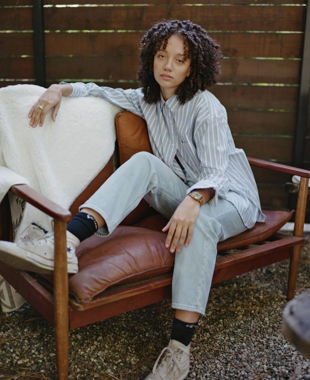 Adult female with curly hair in casual wear poses sitting on a brown leather chair outdoors with a wooden fence in the background.