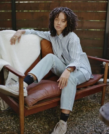 Adult female with curly hair in casual wear poses sitting on a brown leather chair outdoors with a wooden fence in the background.