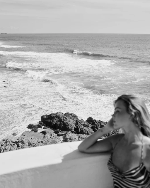 Black and white image of a woman leaning on a wall overlooking the ocean with crashing waves and a rocky shoreline in the distance.