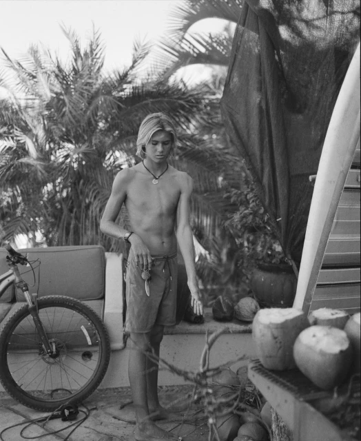 Black and white portrait of a shirtless young man with blond hair standing near a surfboard and coconuts in an outdoor setting.