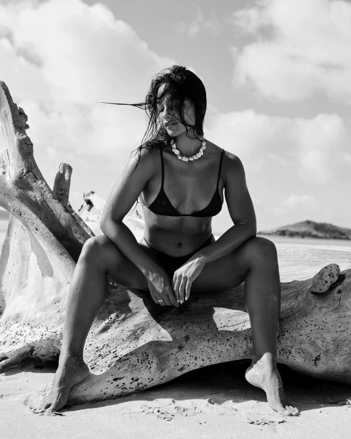 Monochrome portrait of a woman in a bikini and necklace sitting on driftwood on a beach with a cloudy sky in the background.