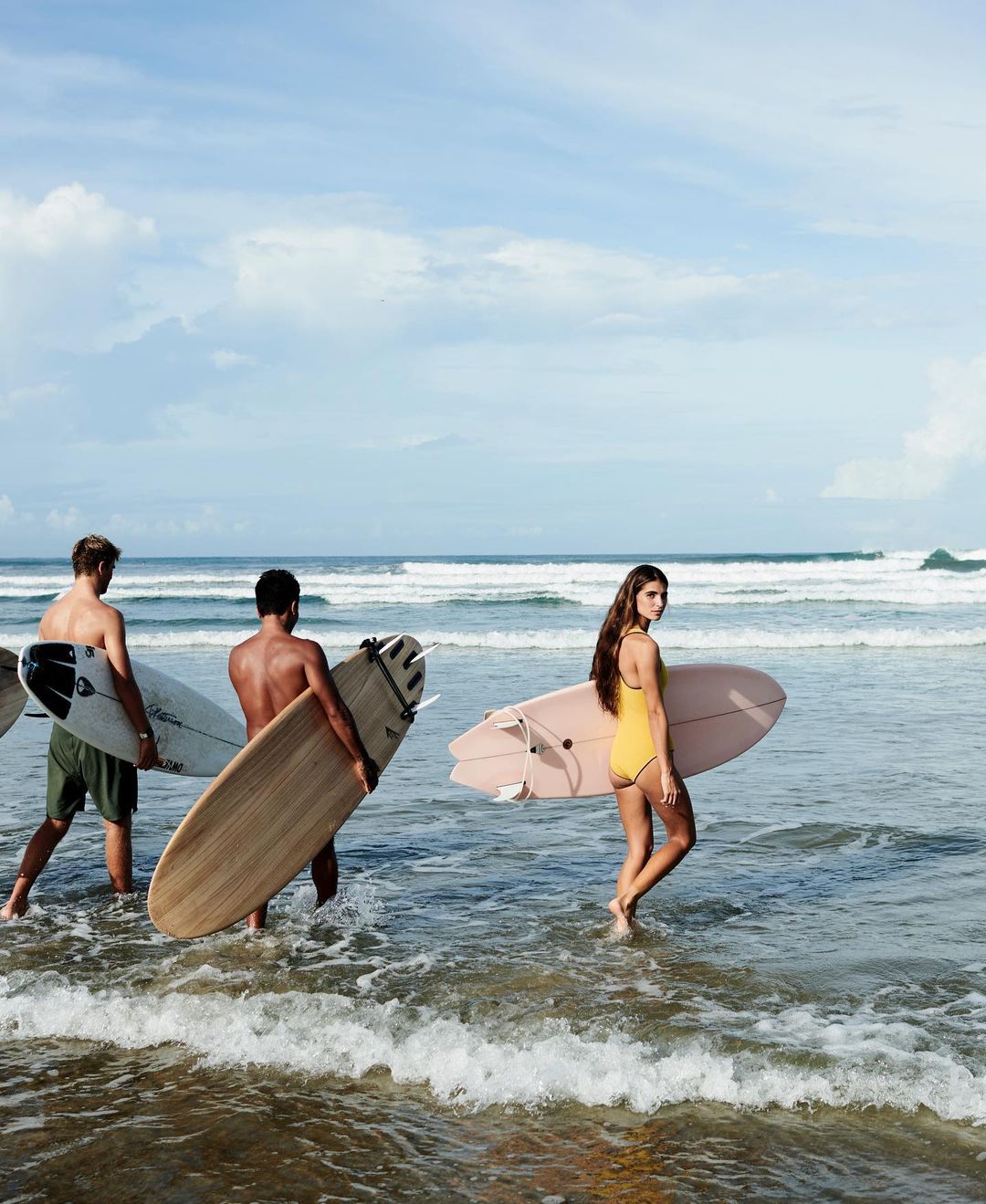 Three people walk in the ocean surf with surfboards. One woman in a yellow swimsuit and two shirtless men carrying surfboards on a sunny day.