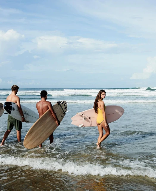 Three people walk in the ocean surf with surfboards. One woman in a yellow swimsuit and two shirtless men carrying surfboards on a sunny day.