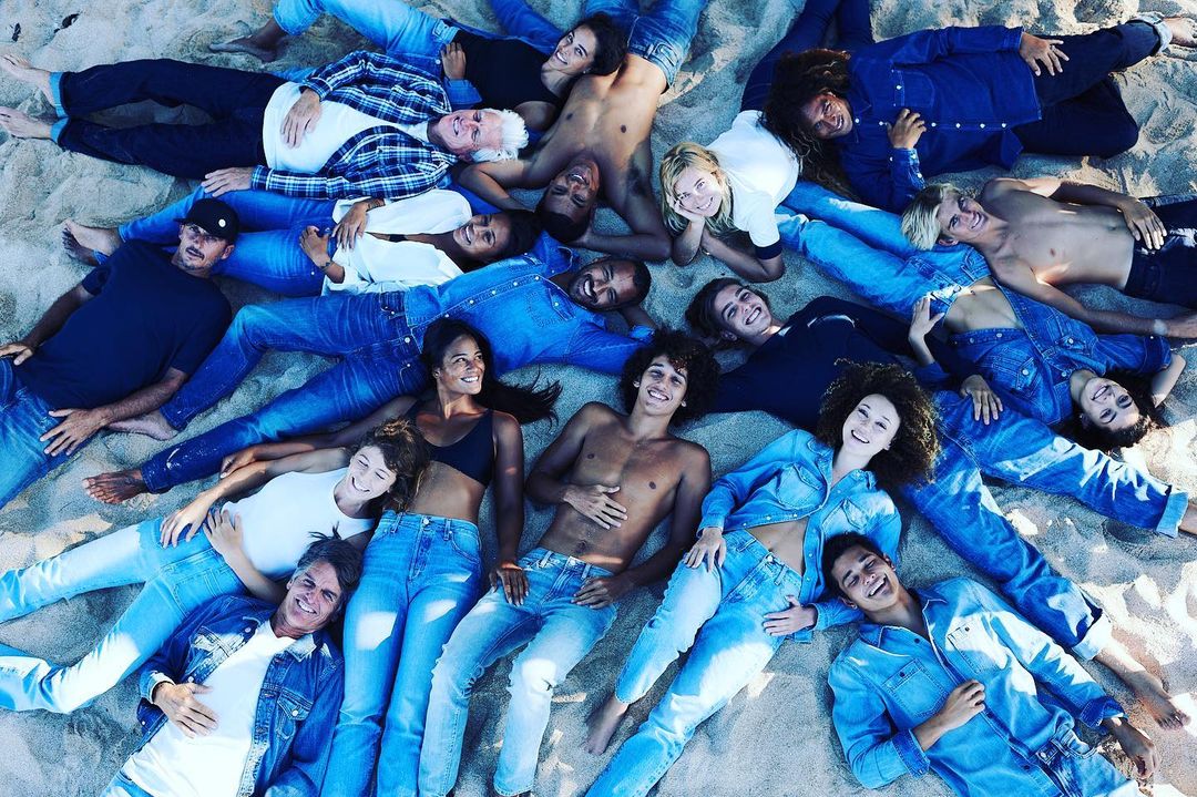 A group of diverse young adults wearing denim lying on sand, top-down view. Casual fashion and beachside setting create a relaxed, youthful vibe.