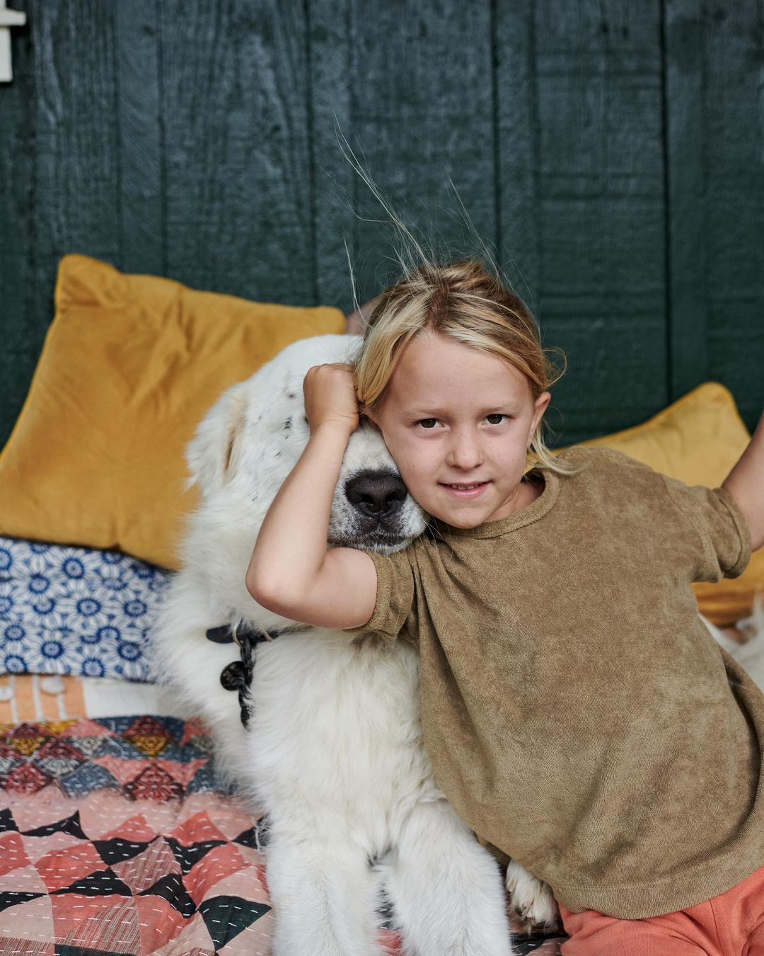 A young blond child hugs a large white fluffy dog while sitting on a bed with colorful quilts and pillows against a dark green wall.