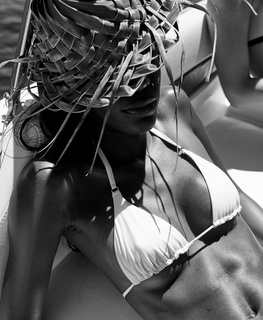 Monochrome shot of an adult female in a bikini, laying down, with a woven hat covering her eyes, on a white surface.