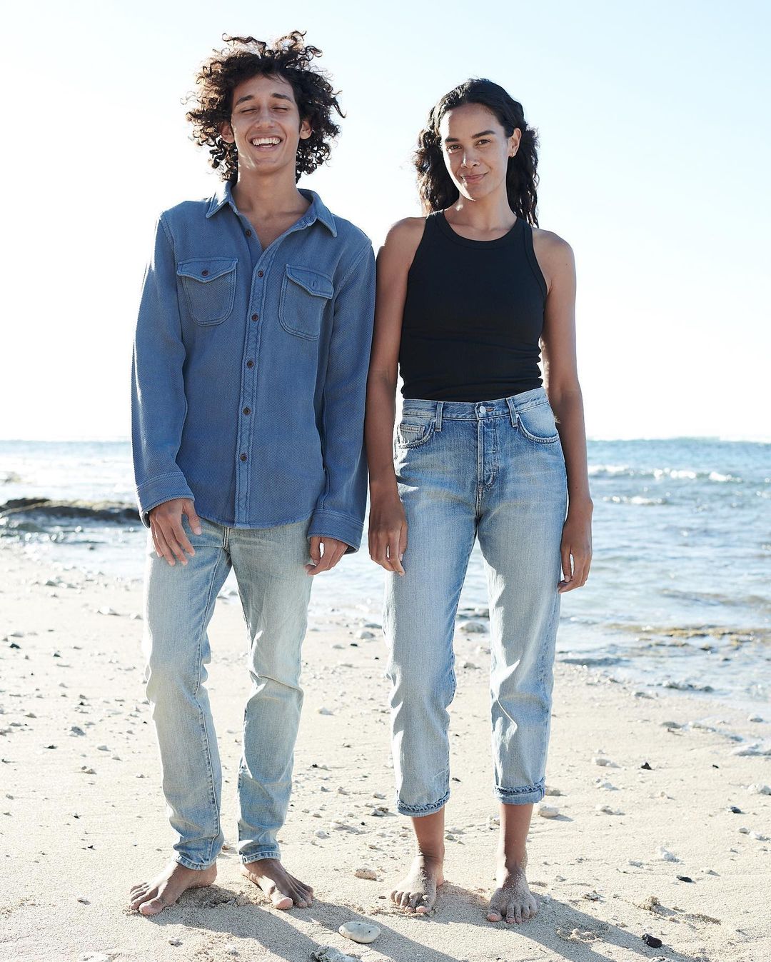 Two people stand on a beach in denim apparel. The male wears a denim shirt and jeans, while the female wears a tank top and jeans.