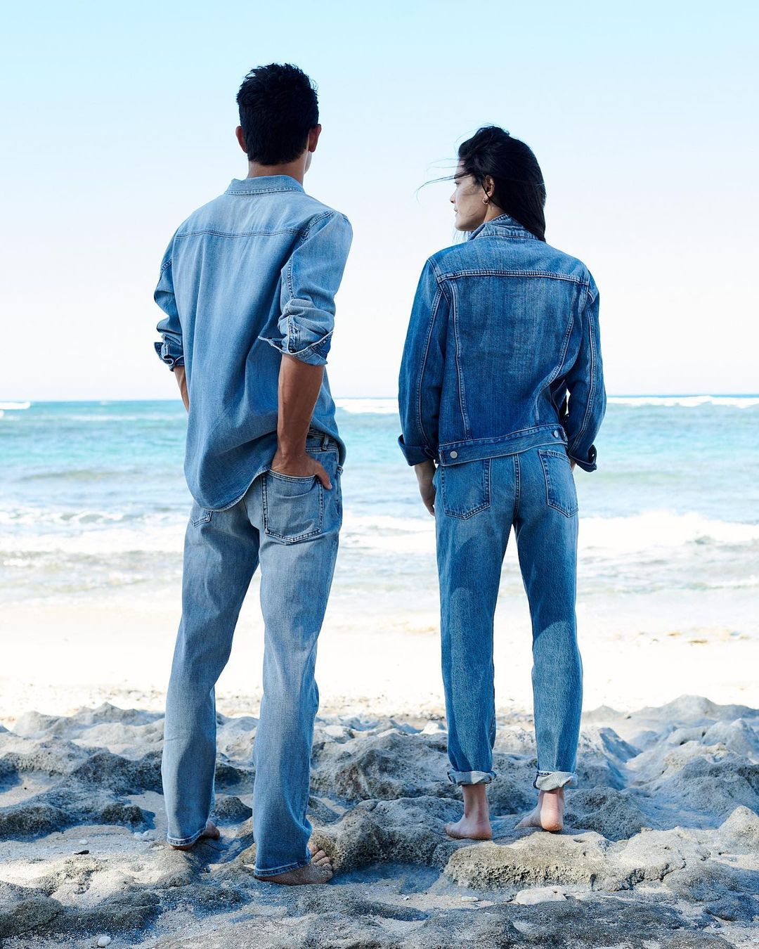 Back view of a stylish couple in denim outfits standing barefoot on a sandy beach, looking out at a turquoise ocean under a bright sky.