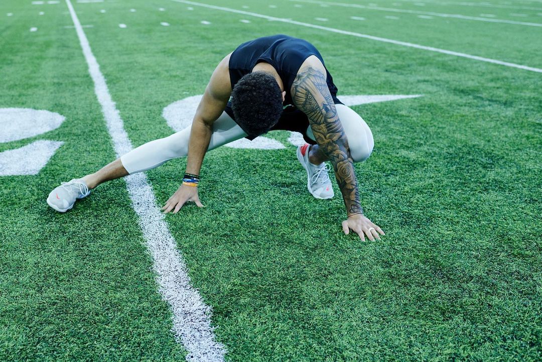 Athlete performs a straddle split stretch on a green sports field with white lines, dressed in athletic wear in an outdoor setting.