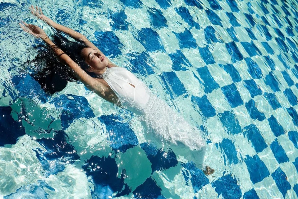 Woman in a flowing white dress floats peacefully in a pool with blue and white checkered tiles, creating a serene and elegant visual.