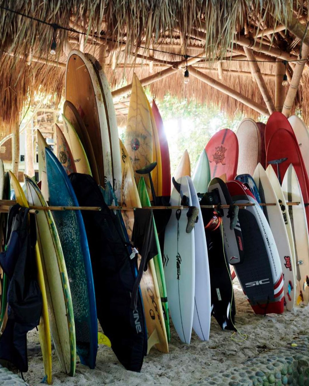 Outdoor surfboard rack filled with an assortment of colorful surfboards on a sandy beach in front of a bamboo structure.