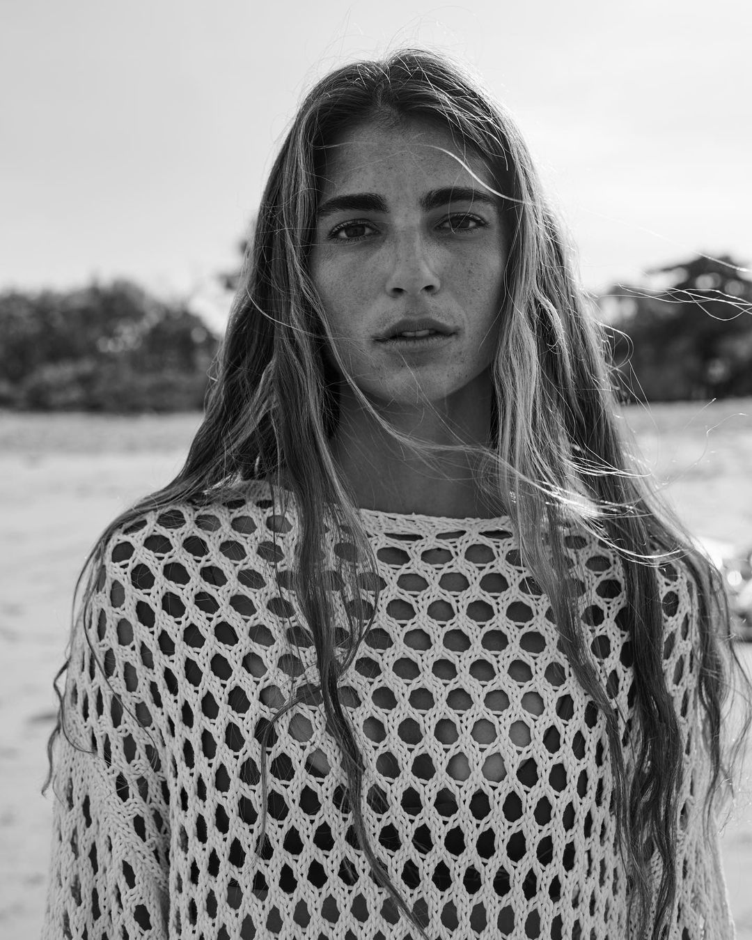 Black and white portrait of a woman with long hair wearing an open-weave knit top outdoors on a beach.