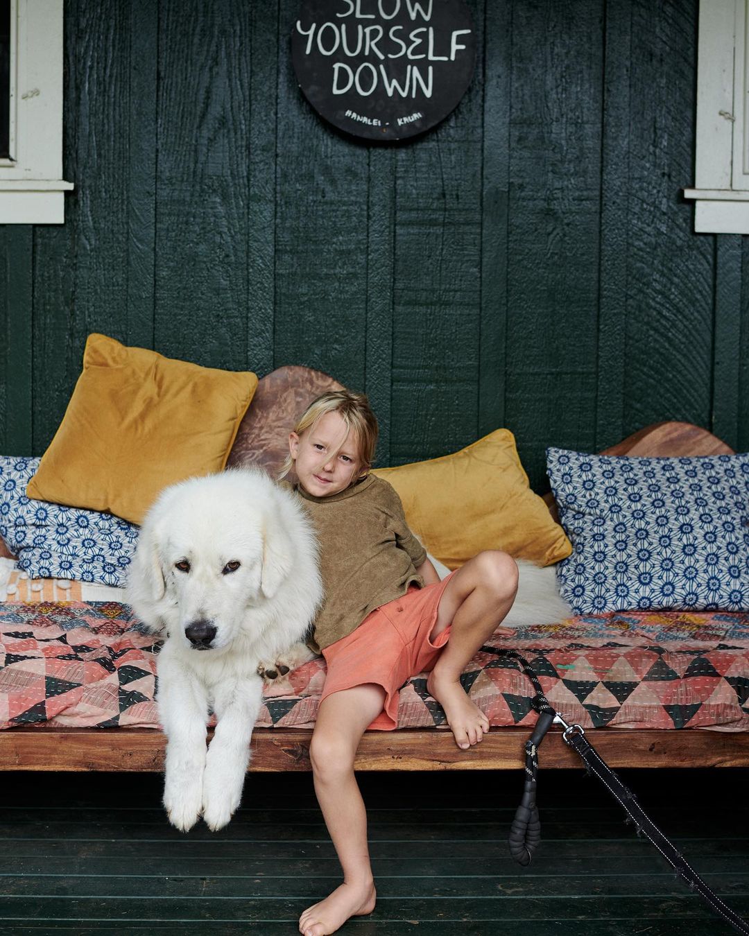 A young boy sits on a wooden bench with patterned cushions next to a fluffy white dog, outside a dark green clapboard wall.
