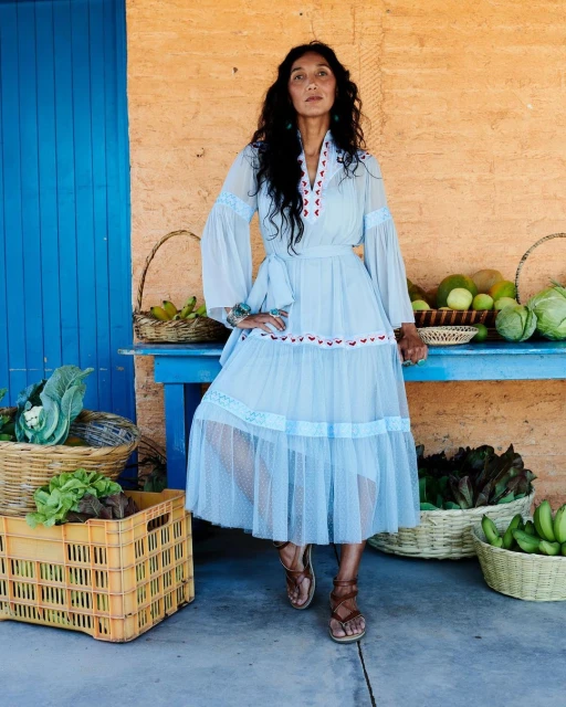 Adult female wearing a long light blue dress with lace trim, standing against a rustic wall with baskets of produce on a blue table.