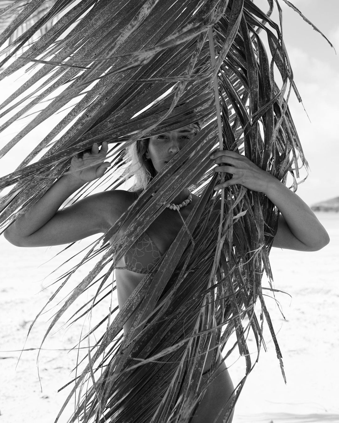 Black and white portrait of an adult female in a bikini partially obscured by palm leaves on a sandy beach beneath a bright sky.