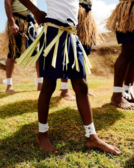 Group of men in traditional clothing with grass skirts and ankle wraps standing on a grassy field in daylight, showcasing culture.
