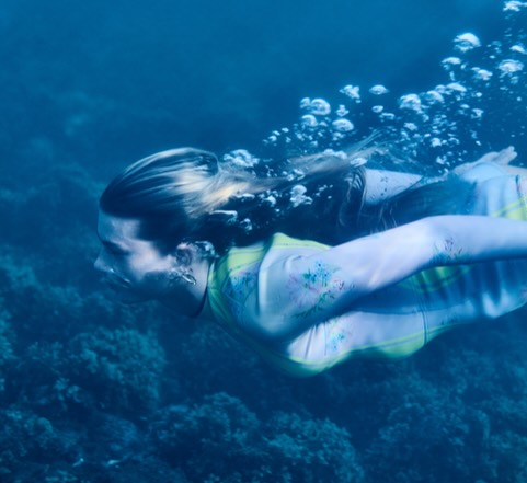 Underwater view of a fair-skinned adult female swimming in the ocean with blue water, coral, and white bubbles.