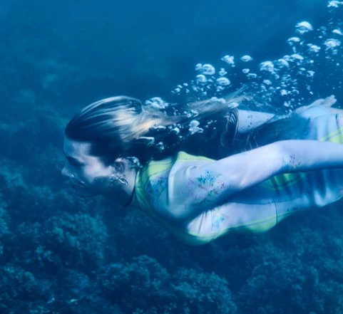 Underwater view of a fair-skinned adult female swimming in the ocean with blue water, coral, and white bubbles.