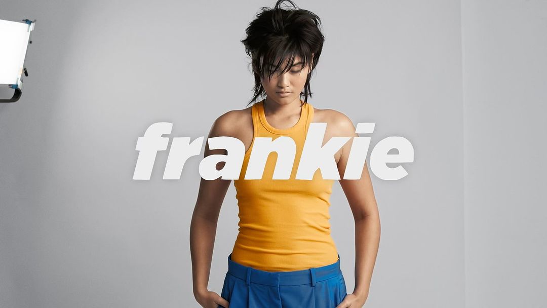 A young woman with a short hairstyle poses in a studio wearing a yellow tank top and blue trousers, under studio lighting.
