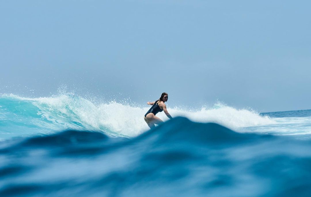 A woman in a dark bathing suit is surfing a barreling wave in clear blue water on a sunny day in an outdoor ocean environment.