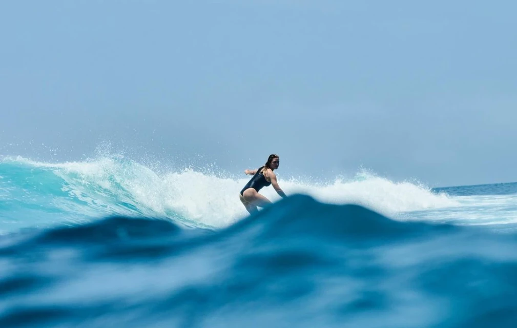 A woman in a dark bathing suit is surfing a barreling wave in clear blue water on a sunny day in an outdoor ocean environment.