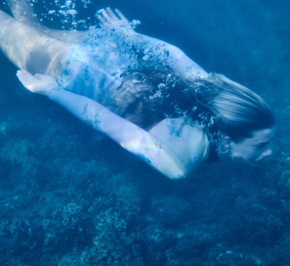 Woman swimming underwater in blue ocean water with sunlight dappling through the surface, coral reef visible on the ocean floor.