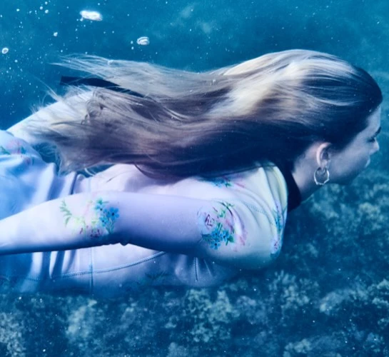 Underwater portrait of a young woman with flowing blonde hair, floral-patterned top, and hoop earrings, swimming gracefully in blue water.