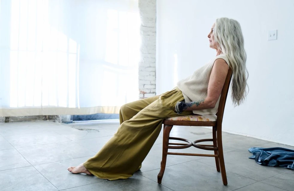 An elderly woman with long gray hair is sitting sideways on a wooden chair in a brightly lit studio, wearing a beige sweater and olive pants.