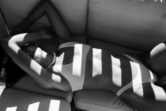 Monochrome image of a woman in a white bikini lying on a couch with dramatic striped shadows across her body by Anne Menke