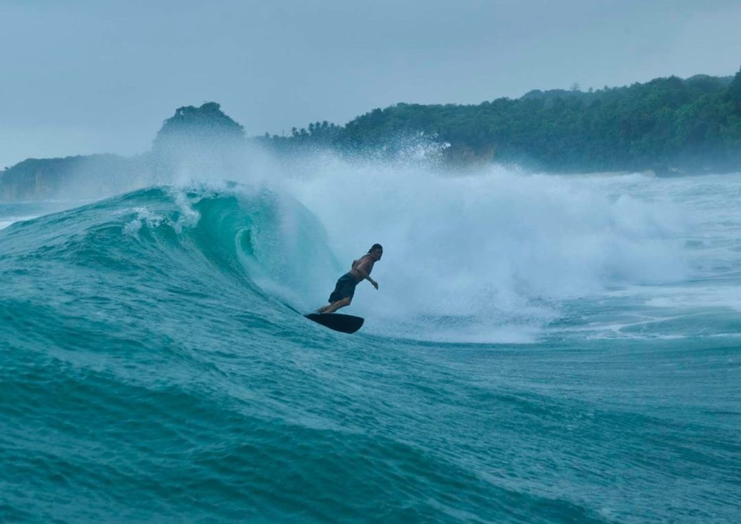 Surfer riding a large wave in the ocean near a tropical island with lush green trees and cloudy skies, showcasing the power of nature.