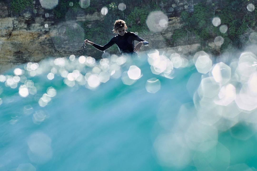 Young surfer in wetsuit stands in clear turquoise water, ocean sparkling with light, rocky cliffs in background.