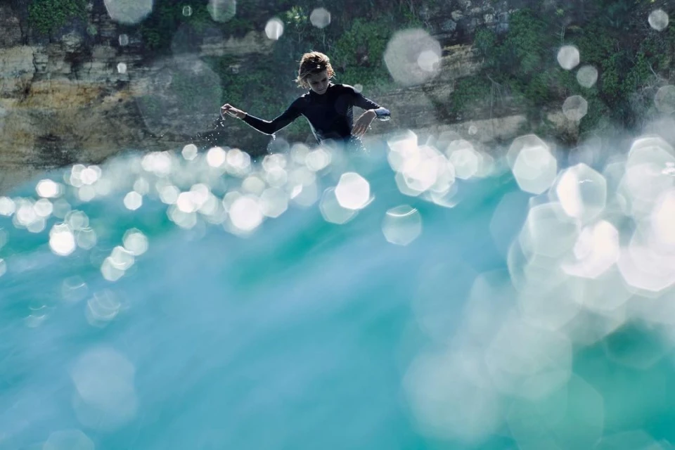Young surfer in wetsuit stands in clear turquoise water, ocean sparkling with light, rocky cliffs in background.