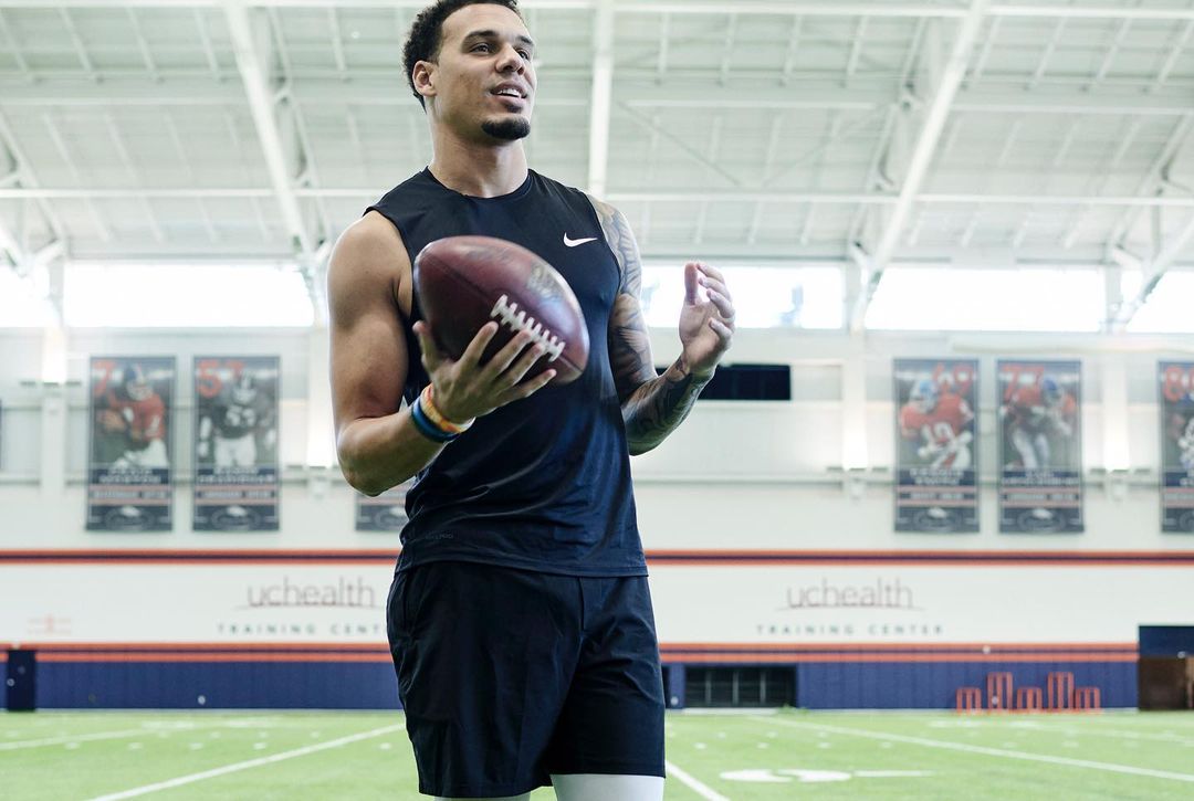 Athletic man in black sportswear holding a football indoors with a large indoor field and a blurred sports banner in the background.