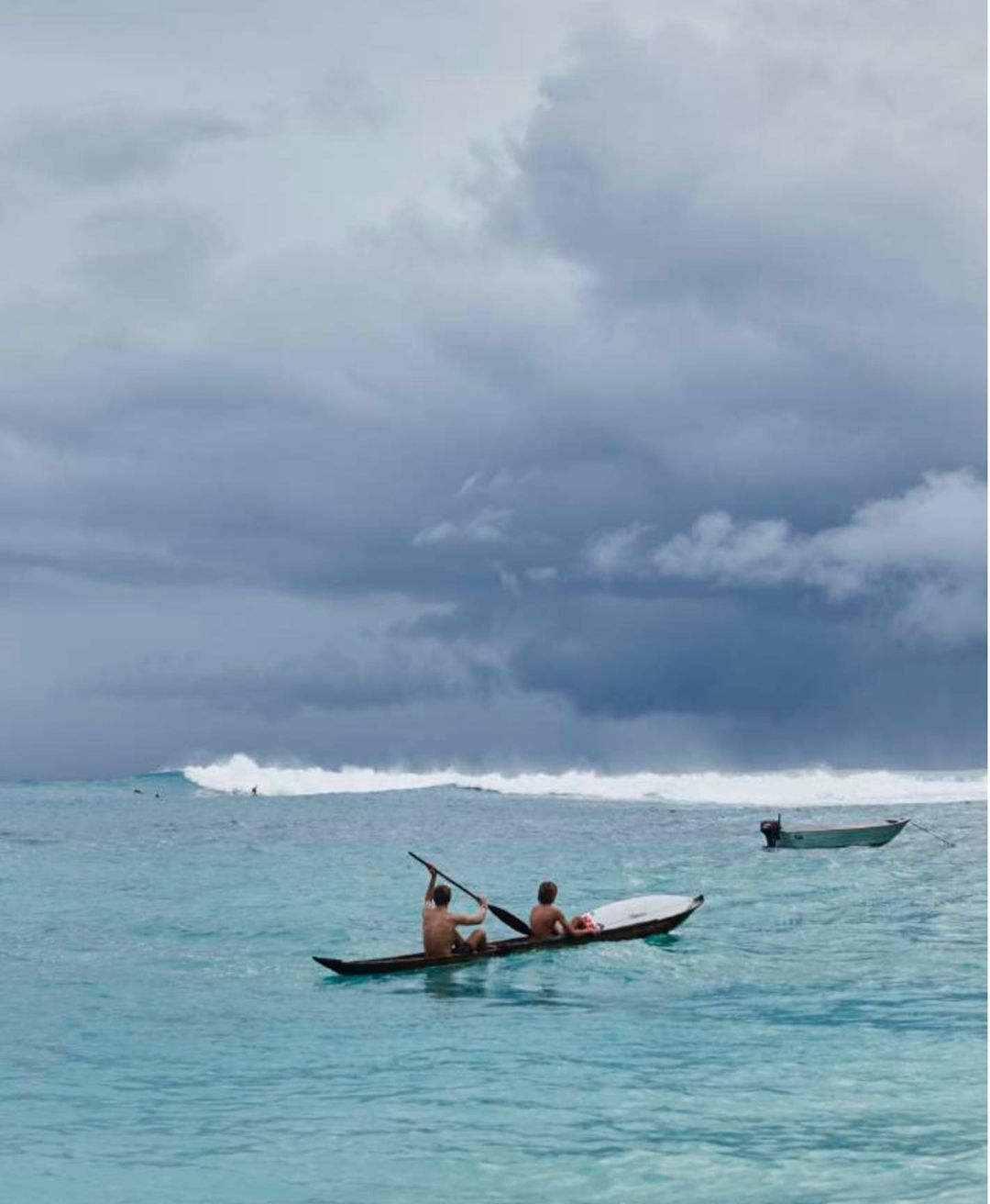 Two men paddle a long, narrow boat in turquoise water under a cloudy sky with a distant wave and another boat visible.
