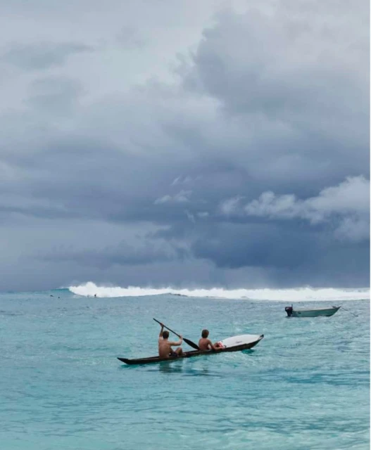 Two men paddle a long, narrow boat in turquoise water under a cloudy sky with a distant wave and another boat visible.
