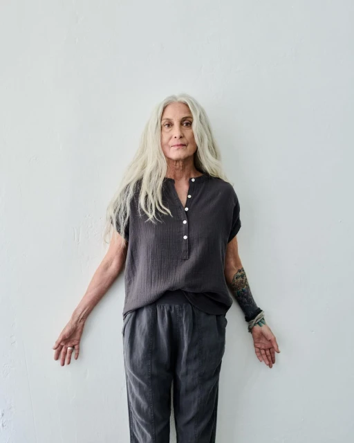 Elegant mature woman with long grey hair posing in casual gray shirt and pants against a white wall in an indoor setting.