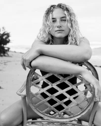 Portrait of a woman with curly blonde hair resting arms on the back of a vintage metal chair on a sandy beach.