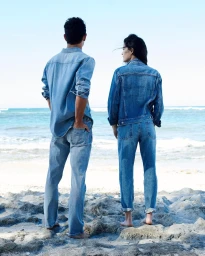 Two models in matching denim shirts and jeans standing barefoot on a rocky shore looking out at the ocean.