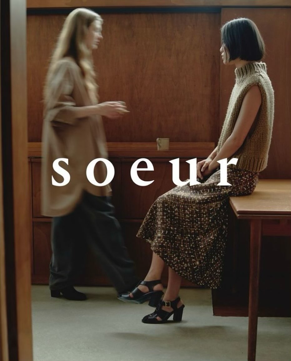 Two women in a naturally lit indoor setting, one seated on a wooden table wearing a midi skirt, facing the other.